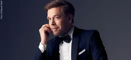 Person in elegantem Anzug mit Fliege schaut nachdenklich mit leichtem Lächeln in die Ferne.