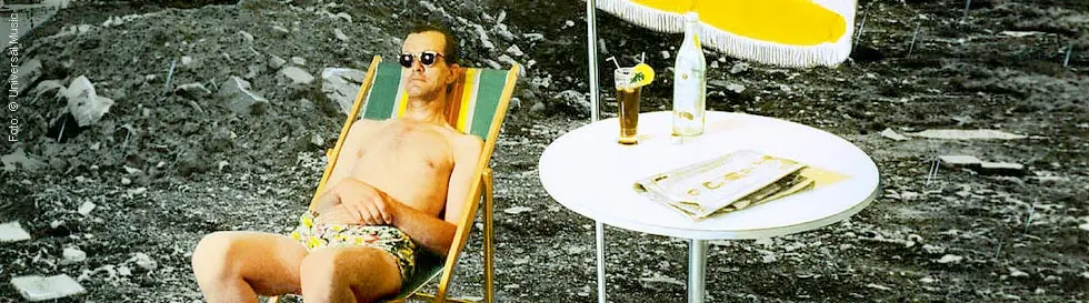 Ein Mann in Badehose und Sonnenbrille liegt auf einem Liegestuhl. Daneben ein Tisch mit Getränk und Zeitung. Hintergrund ist eine Baustelle.