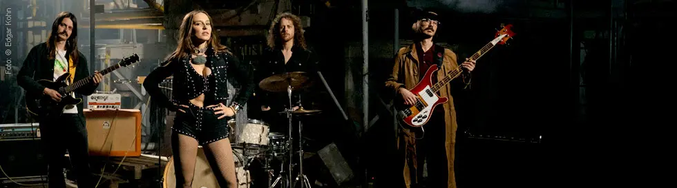 Vier Musiker stehen in einer industriellen Umgebung. Eine Frau steht vorn in roten Stiefeln, umgeben von drei Männern mit Instrumenten.