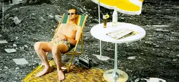 Ein Mann in Badehose und Sonnenbrille liegt auf einem Liegestuhl. Daneben ein Tisch mit Getränk und Zeitung. Hintergrund ist eine Baustelle.