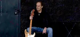 Ein Mann mit Kreuzkette sitzt und hält eine E-Gitarre, links eine Tür.