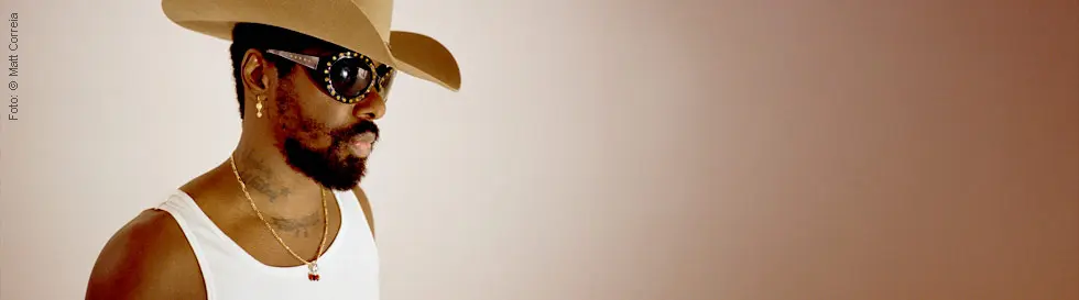 Ein Mann mit Cowboyhut, Sonnenbrille und Ohrringen, trägt ein weißes Shirt und hat einen Bart.