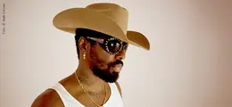Ein Mann mit Cowboyhut, Sonnenbrille und Ohrringen, trägt ein weißes Shirt und hat einen Bart.