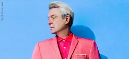Ein Mann mit grauem Haar trägt einen rosa Anzug und Hemd, vor blauem Hintergrund, blickt zur Seite.