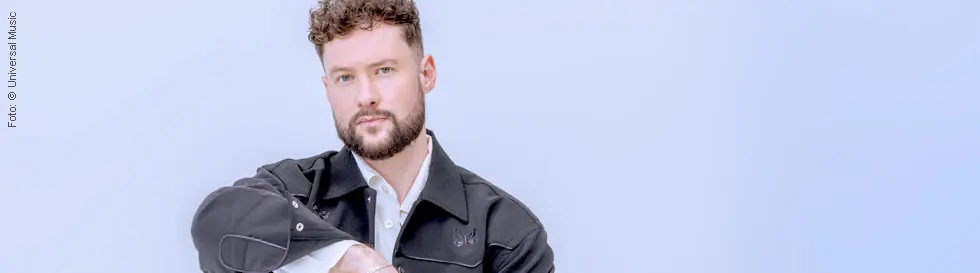 Ein Mann mit lockigem Haar und Bart schaut ernst. Er trägt eine dunkle Jacke und sitzt vor hellem Hintergrund.
