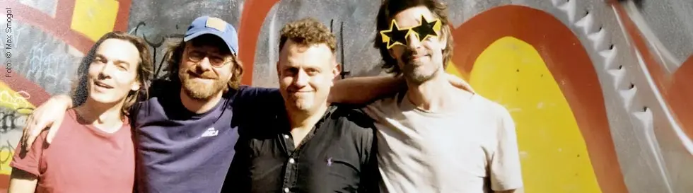 Vier Männer lachen in Freizeitkleidung vor bunter Wand. Einer trägt eine sternförmige Sonnenbrille.