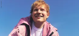 Eine Person mit rötlichen Haaren lächelt nach oben. Sie trägt eine rosa Jacke und ein weißes T-Shirt vor blauem Himmel.