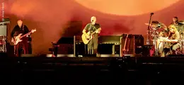 Vier Musiker auf einer Bühne, einer spielt Gitarre, ein anderer sitzt am Schlagzeug, warme Beleuchtung.