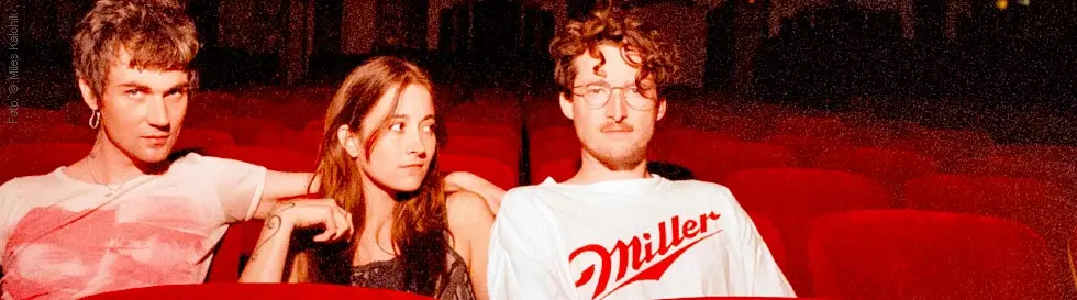 Zu sehen sind drei Personen in einem Theater. Rechts trägt eine Person ein weißes T-Shirt.