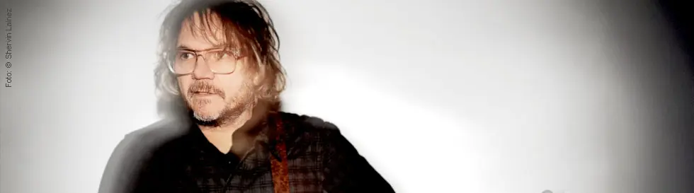 Der Mann hat mittellange Haare, trägt eine Brille und ein kariertes Hemd. Der Hintergrund ist unscharf.