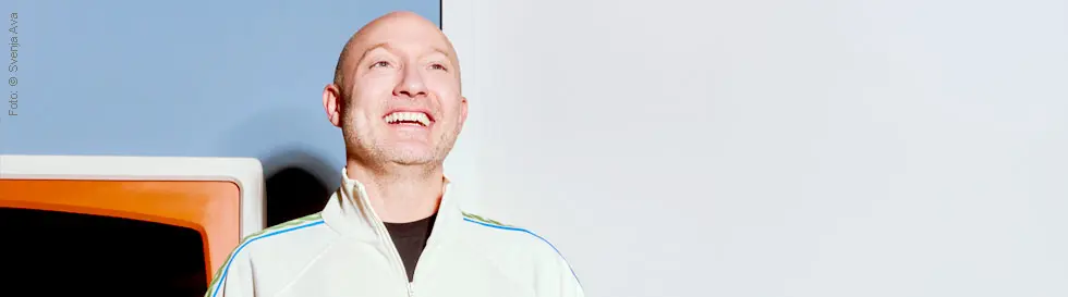 Person in weißer Jacke lächelt breit; Hintergrund orange und hellblau.