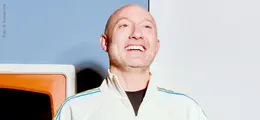 Person in weißer Jacke lächelt breit; Hintergrund orange und hellblau.
