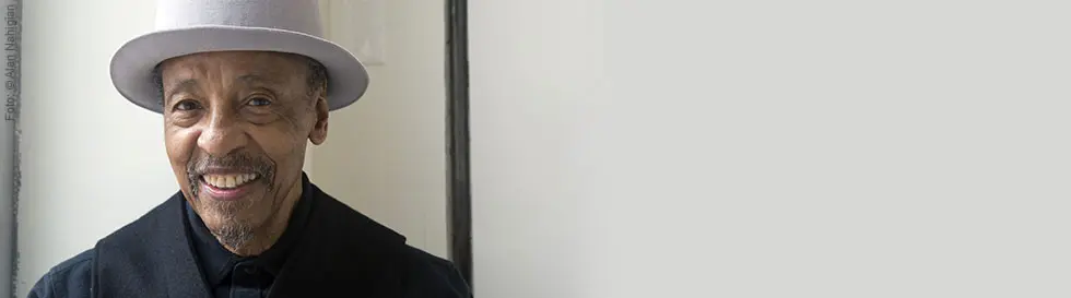 Ein lächelnder Mann mit grauem Hut und dunkler Kleidung vor hellem Hintergrund.