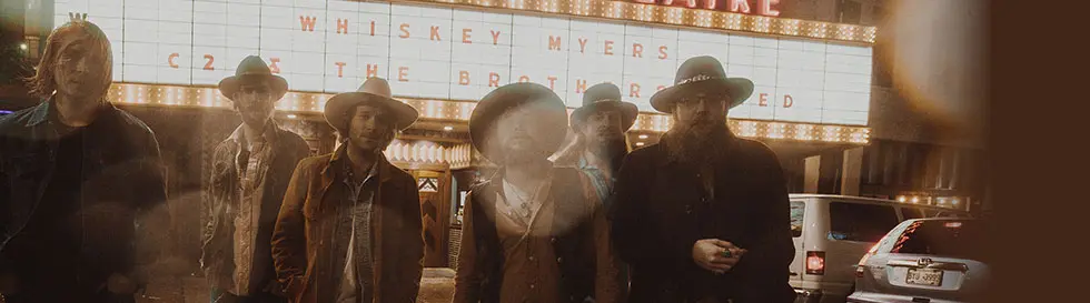 Auf einem Schild steht: "Whiskey Myers". Sechs Männer mit Hüten und Jacken stehen davor.