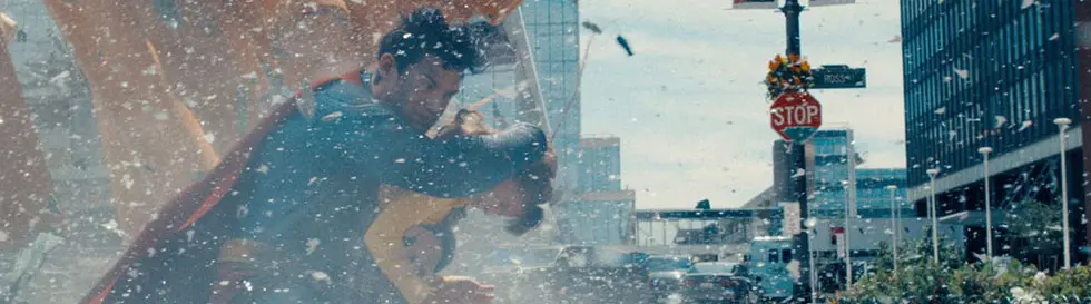 Ein Mann in blauem Anzug mit rotem Umhang schützt ein Kind vor Trümmern auf der Straße. Im Hintergrund ist ein Stoppschild.