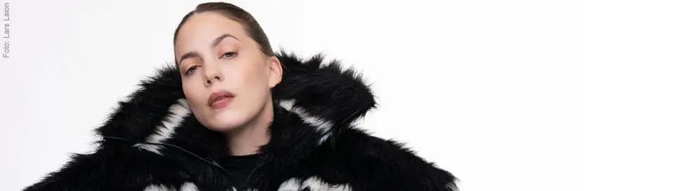 Frau in schwarzem, pelzigem Mantel mit weißen Akzenten, hält ihre Arme vor sich. Hintergrund weiß.