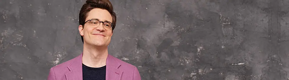 Ein Mann mit Brille und lila Sakko vor grauem Hintergrund.