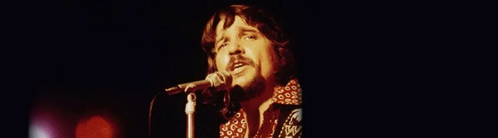 Ein Mann mit Bart singt in ein Mikrofon, trägt ein gemustertes Hemd. Der Hintergrund ist dunkel.