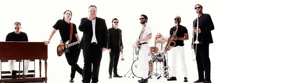 Mehrere Männer in formeller und Freizeitkleidung, mit Musikinstrumenten vor weißem Hintergrund.