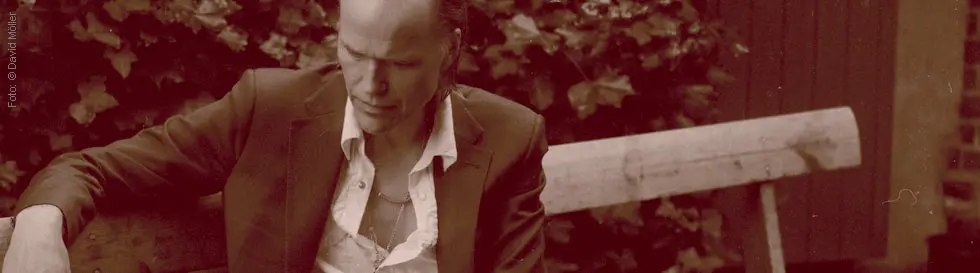 Ein Mann in einem Anzug sitzt auf einer Holzbank, umgeben von Efeu in einem sepiafarbenen Ton.