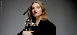 Eine Frau mit blondem Haar hält ein Saxofon, sie trägt schwarze Kleidung. Der Hintergrund ist grau.
