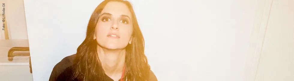 Eine Frau mit Gitarre in entspannter Pose, trägt ein buntes T-Shirt.