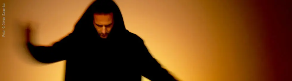 Eine Person mit Kapuze, Kopf gesenkt, in einer unscharfen Bewegung vor orangefarbenem Hintergrund.