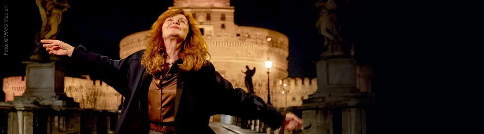 Eine Person steht mit ausgebreiteten Armen, im Hintergrund eine beleuchtete Festung.