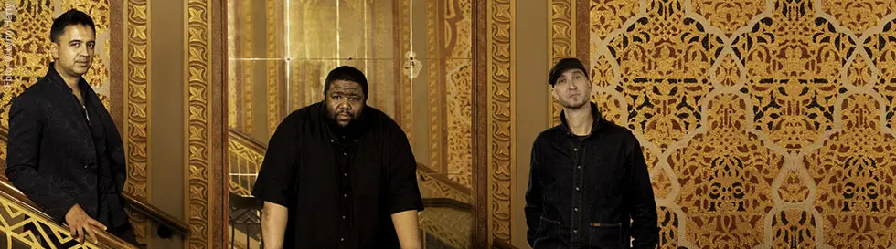 Drei Männer in dunkler Kleidung vor einer reich verzierten, gold- und rotfarbenen Wand und Treppe.