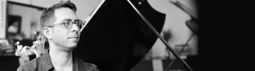 Eine Person mit Brille sitzt vor einem Flügel, im Hintergrund sind unscharfe Elemente zu erkennen.