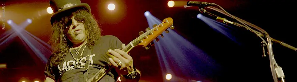 Ein Musiker mit Zylinder spielt Gitarre auf einer Bühne. Im Hintergrund bunte Lichter.