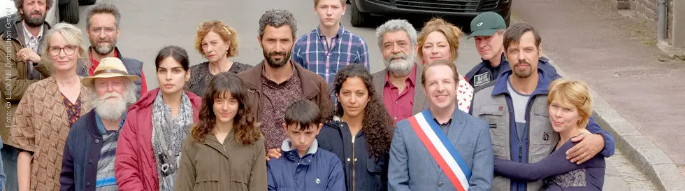 Eine Gruppe von 17 Menschen steht in einer Straße zusammen. Eine Person trägt eine Schärpe in den französischen Nationalfarben.