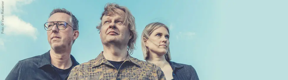 Drei Personen schauen in die Ferne, vor blauem Himmel. Links ein Mann mit Brille, in der Mitte ein weiterer Mann, rechts eine Frau.