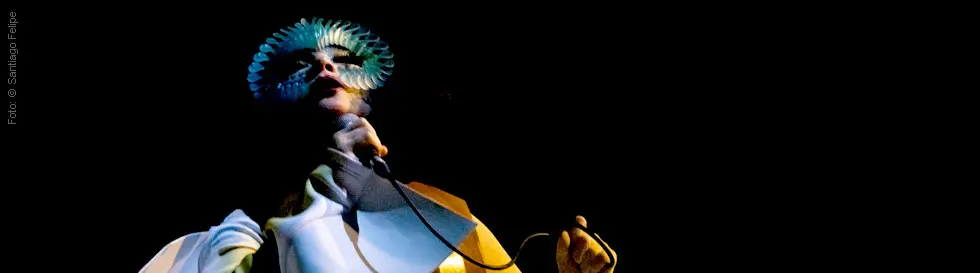 Person im futuristischen Kostüm, hält ein Mikrofon, Hut aus blauen Federn.