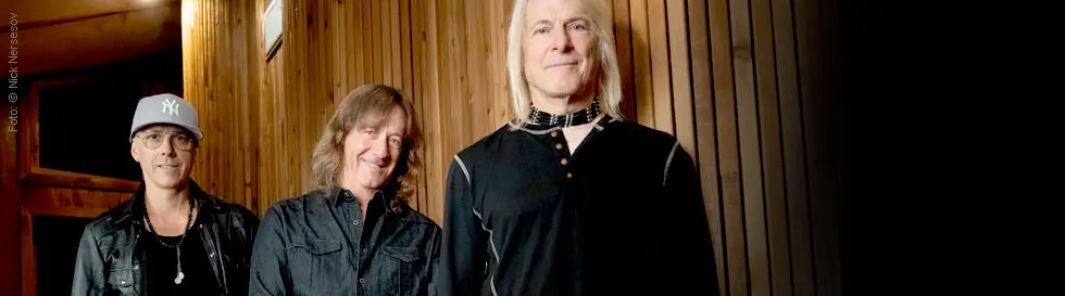 Drei Männer in einem holzgetäfelten Raum. Einer trägt eine Baseballkappe, ein anderer eine schwarze Jacke.