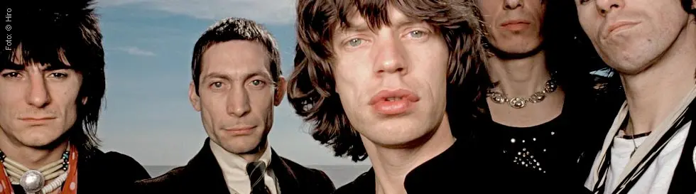 Es sind fünf Männer mit langen Haaren und unterschiedlichen Ausdrücken vor einem blauen Himmel zu sehen.