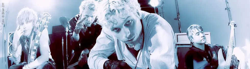 Vier Personen in auffälliger Kleidung mit Instrumenten, eine hält ein Mikrofon vor sich.