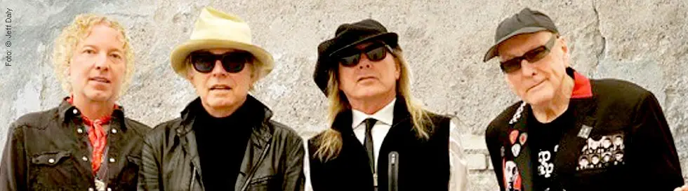 Vier Männer in modischer Kleidung vor einer strukturierten Wand, zwei mit Hut und Sonnenbrille.