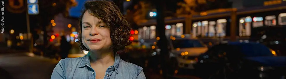 Eine Frau mit lockigem Haar lächelt, trägt ein jeansblaues Hemd, steht abends auf einer belebten Straße mit Lichtern.
