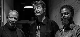 Drei Männer stehen nebeneinander in einem dunklen Raum. Sie schauen ernst, sind unterschiedlich gekleidet und haben unterschiedliche Frisuren.