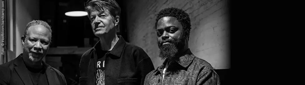 Drei Männer stehen nebeneinander in einem dunklen Raum. Sie schauen ernst, sind unterschiedlich gekleidet und haben unterschiedliche Frisuren.