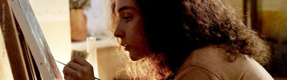 Eine Frau malt konzentriert auf einer Leinwand mit einem Pinsel, im Hintergrund sind unscharfe Blumen.