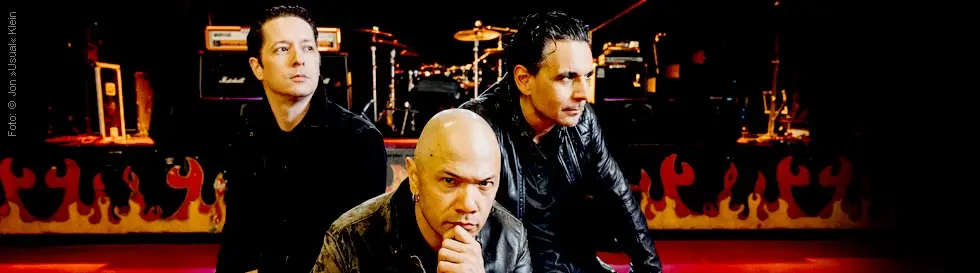 Drei Männer in Lederjacken posieren vor einer Bühne mit Schlagzeug und Verstärkern, im Hintergrund sind Flammenmotive.