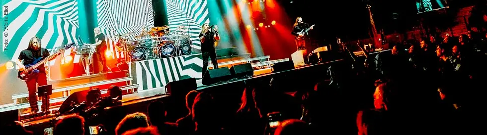 Fünf Musiker auf einer auffällig beleuchteten Bühne mit dynamischem Streifenmuster im Hintergrund. Mehrere Zuschauer mit Handys.