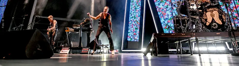 Drei Musiker auf einer Bühne, ein Sänger mit Mikrofon, ein Gitarrist und ein Keyboarder vor lebhafter Lichtshow.