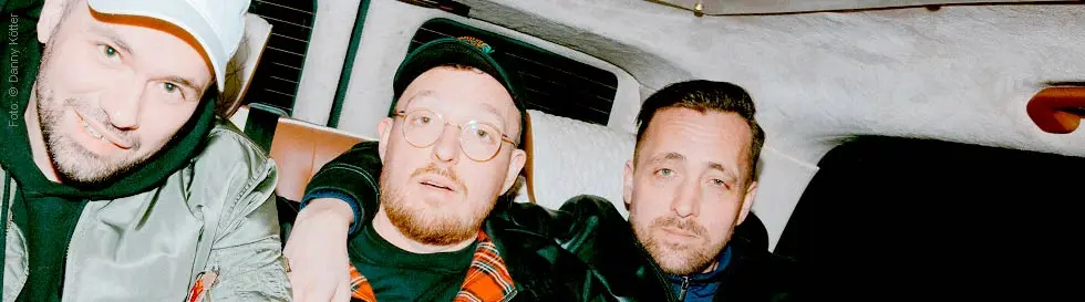 Drei Männer sitzen eng nebeneinander auf einem Rücksitz. Der linke trägt eine grüne Jacke und eine Kappe.