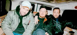 Drei Männer sitzen eng nebeneinander auf einem Rücksitz. Der linke trägt eine grüne Jacke und eine Kappe.