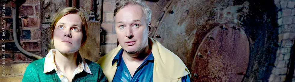 Ein Mann und eine Frau stehen vor einer alten Backsteinwand, schauen aufmerksam geradeaus, die Frau trägt einen grünen Pullover.