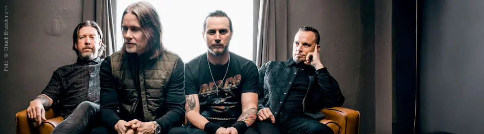 Vier Männer sitzen auf einem Sofa. Einer trägt ein T-Shirt mit Bandlogo. Die Atmosphäre ist entspannt und nachdenklich.