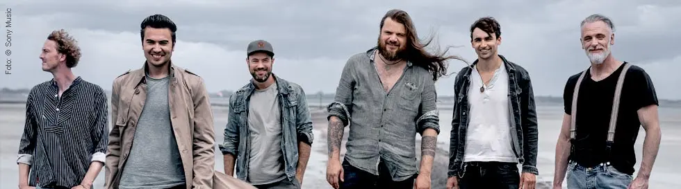 Sechs lächelnde Männer stehen lässig auf einem Steinweg am Wasser. Der Himmel ist bewölkt.